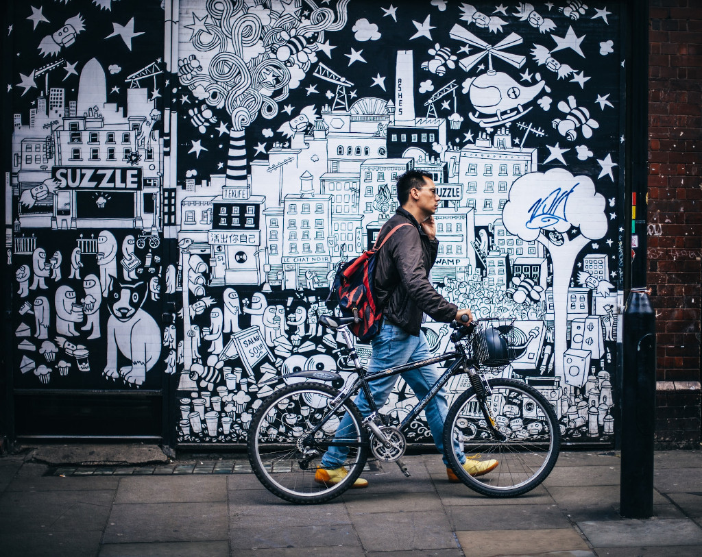 un homme marchant sur un trottoir un vélo dans une main et un téléphone dans l'autre. Le mur derrière représente une ville